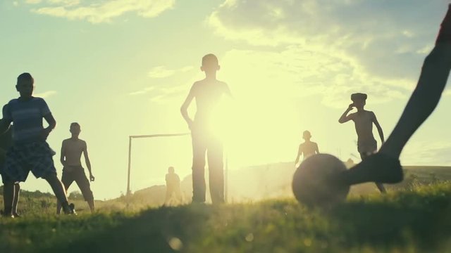 Simple poor farmer boys playing football on the background of a bright sunset in summer hot day. Slow motion.