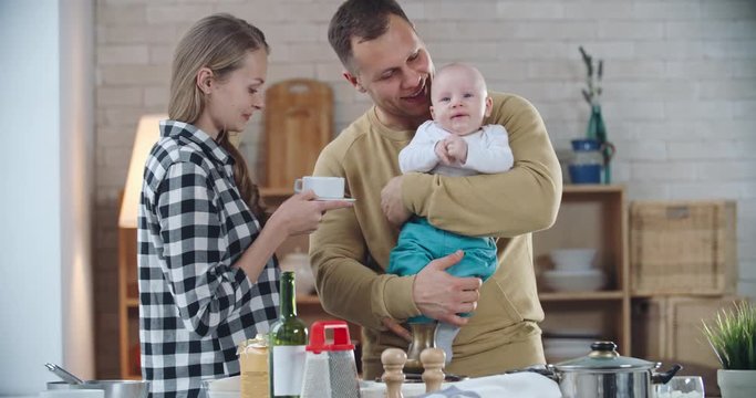 Beautiful family spending morning together: man standing with baby and kissing his beautiful wife holding coffee