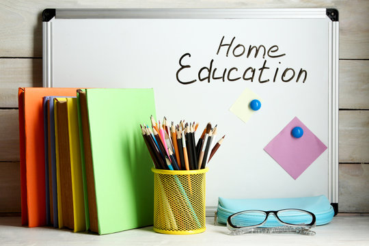 Board with the inscription "home educktion" near the pencil box with school equipment and stack of books on white wooden background