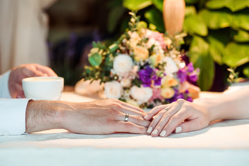 Man and woman's hands with espousal rings, coffee, wedding. Closeup of bride and groom holding hands in cafe. Wedding rings on their fingers.