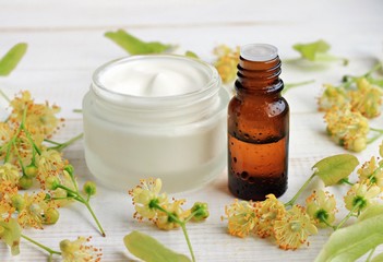 Jar of white cosmetic facial cream, essential oil bottle, linden (tilia) blossom flowers. Holistic herbal skincare. 