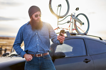 bearded traveler using smart phone in front of car with bike rack on road trip in nevada