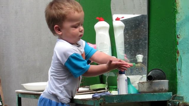 The child washes his hands alone in an old sink with liquid soap and water in the yard of the old house without the help of parents summer day