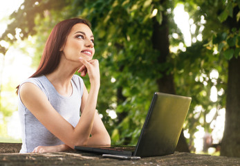 Beautiful young woman sitting in the park, using a laptop, thinking