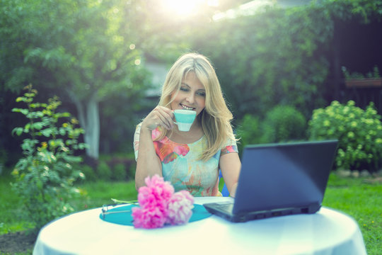 Красивая блондинка пьет кофе и смотрит в ноутбук за столом в саду