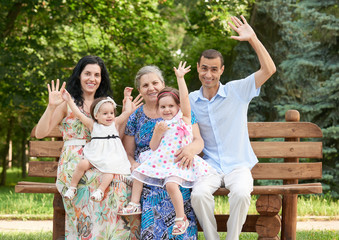 Naklejka premium big family sit on wooden bench in city park and waving, summer season, child, parent and grandmother, group of five people