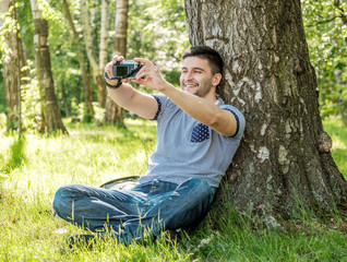 Portrait of young hipster man with camera outdoors. Young male photographer making selfie on summer day.