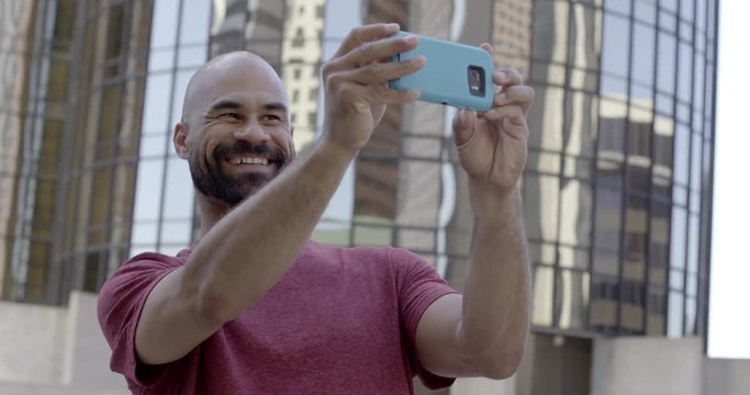 Handsome mixed race man takes selfies on his cellphone while standing in front of large buildings in Downtown Los Angeles.  Medium shot, recorded in slow motion at 60fps.