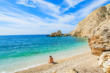 Young attractive woman sitting on beach in swimsuit and looking at turquoise sea water in Petani bay, Kefalonia island, Greece