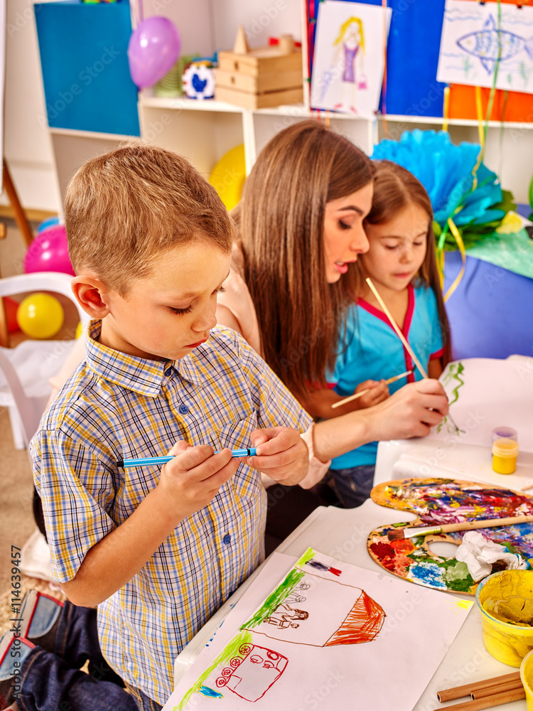 Wall mural Children with teacher woman painting on paper at table in kindergarten .