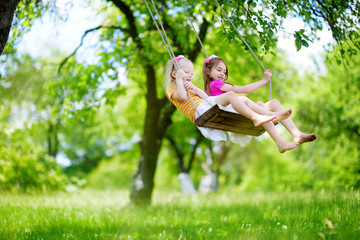 Two cute little sisters having fun on a swing together in beautiful summer garden
