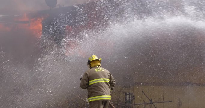 Slow-motion view of a fireman with a hose walking toward a burning house while water droplets pour in from right of frame
