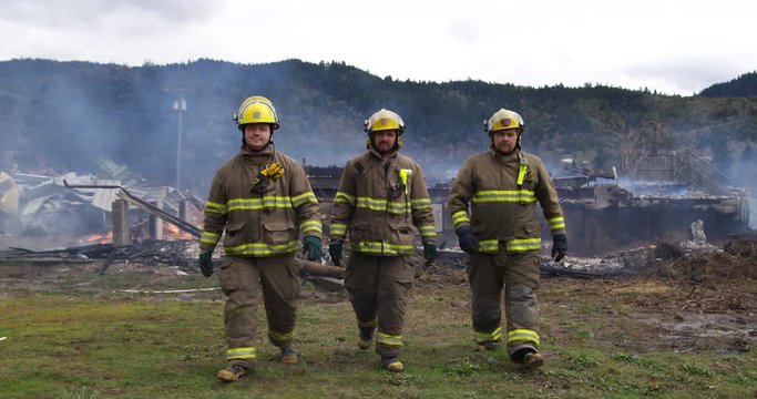 Slow-motion shot of three firemen walking towards the camera with a smoldering structure in the background