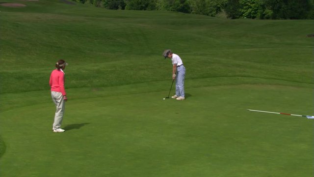 Woman golfer putting as companion helps line up shot, ball rolls short of hole