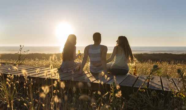 Friends sitting on boardwalk at the beach, drinking beer at sunset