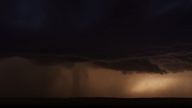 Lightning flashes out of a dark thunderstorm and a curtain of rain partially hides sunset glow above dark hills, time lapse