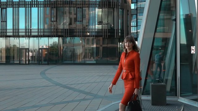 Slow motion. Young sexy businesswoman in red suit with ladies bag and glasses exit from modern business centre building rolling door. Look at the camera. Middle shot. Teal and orange.