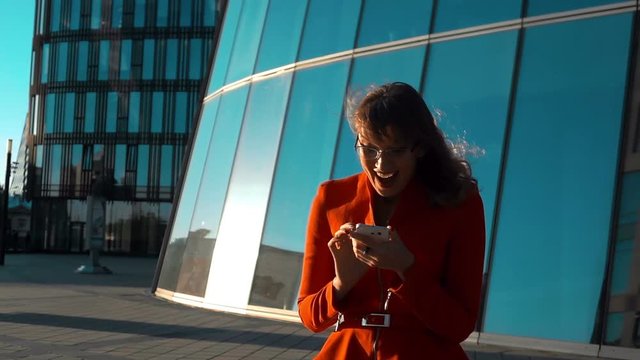 Slow motion. Euphoric sexy woman in red suit and spectacles watching her smartphone read message, rejoice winner with glossy modern business centre buiding at the background. Middle shot.