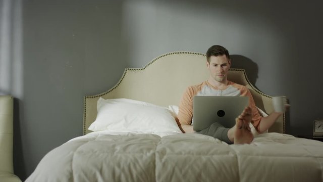 Medium shot of man with coffee using laptop on bed / Cedar Hills, Utah, United States