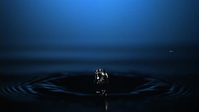 Close-up ultra-slow motion drop falling with high rebound onto glowing blue water