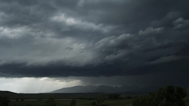 Gray clouds hurry above a darker more static layer as rain falls on mountains and valley, time lapse