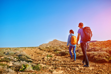 Naklejka premium couple of travelers with backpack walking on the mountain trail against sea and blue sky at early morning. Balos beach on background, Crete, Greece