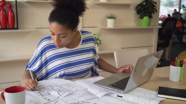 Professional young designer checking plan with electronical version on computer. African mixed race female looking on screen laptop and test plan of new building. girl doing her work at the