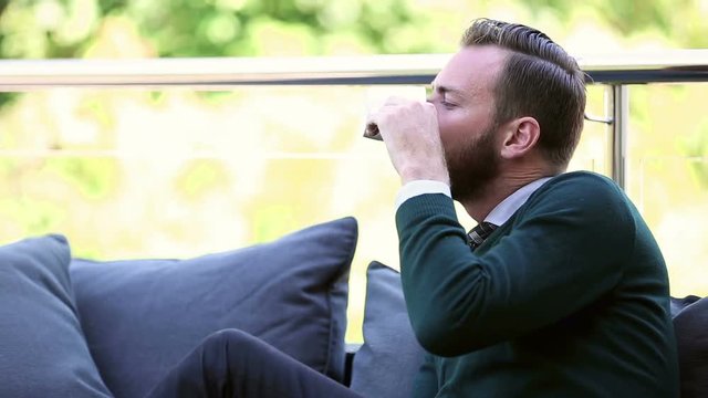 Man wearing a green pullover sitting down outside in a lounge sofa drinking from a mug. Looking at camera on a sunny day.