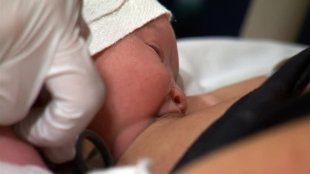 Close-up face of baby sucking at mother's nipple; nurse checking baby with stethoscope