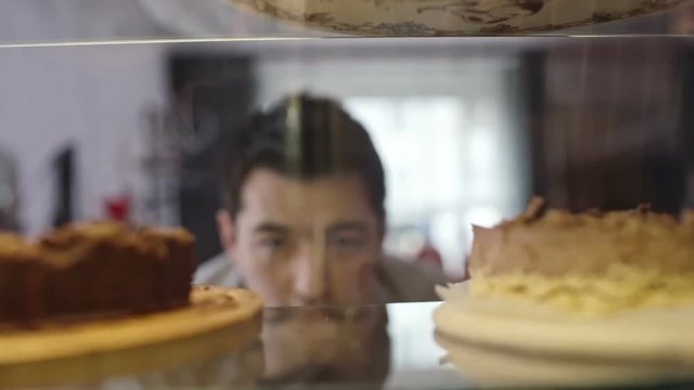 Through glass view of young Asian man choosing cakes at cafe dessert display, slow motion closeup shot on Sony NEX 700