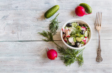 Healthy diet food: vegan vegetable salad with fresh cucumbers, radish, green onion, black olives, dill and feta cheese. In white ceramic bowl on light wooden background.