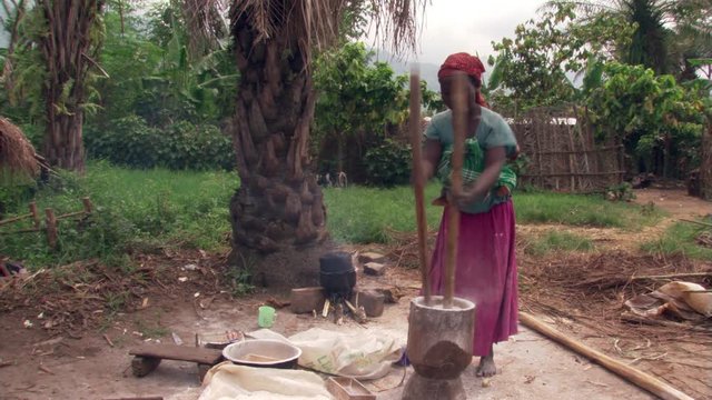 Ugandan woman pounding grain in mortar, baby in sling on her back