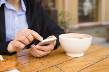 Woman with smartphone sitting in an outdoor cafe and drinking coffee. Breakfast before working day. Cappuccino and croissant on a wooden table. Girl in the cafe.