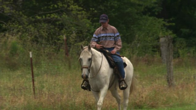 Man riding a gray horse slowly through dry grass toward the camera at an angle