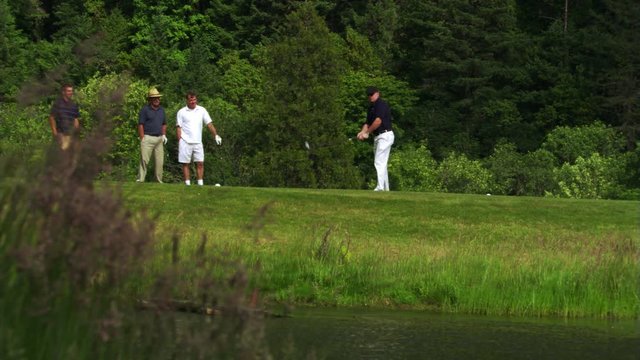 Foursome at water hazard, one golfer hitting ball into water