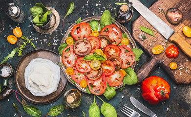 Caprese tomatoes mozzarella salad  on dark rustic background with oil, balsamic vinegar, cutting board and ingredients, top view