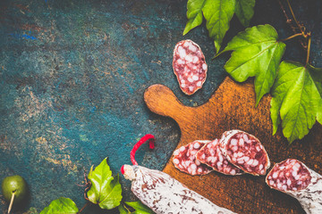 Traditional stick of salami sausage on rustic cutting board on dark vintage background, top view, place for text. Italian food background for menu or recipes .