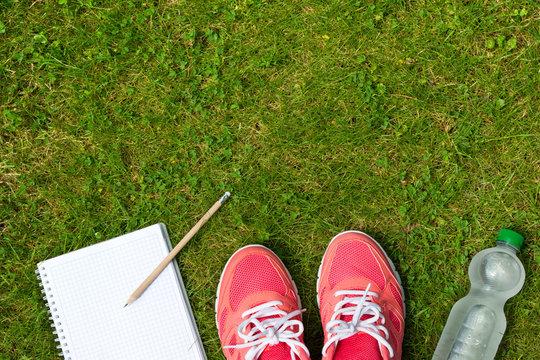 Fitness concept, pink sneakers, notebook with pencil and bottle of cold water on green grass, top view