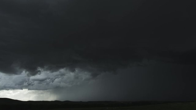 Turbulent supercell clouds pierced by a lightning flash roll above low hills, time lapse