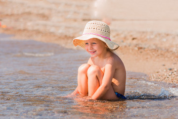 laughing joyful girl in a hat sitting on the beach and playing with sand and water on a sunny day