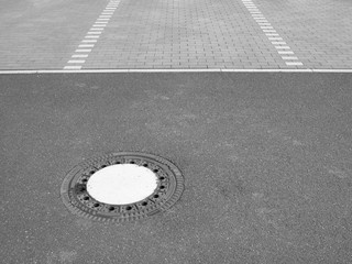 Runder Kanaldeckel in grauen Asphalt auf einem Parkplatz mit Verbundpflaster einer Fabrik im...