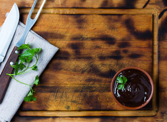 Barbecue sauce in clay bowl, meat fork, knife, cutting board.