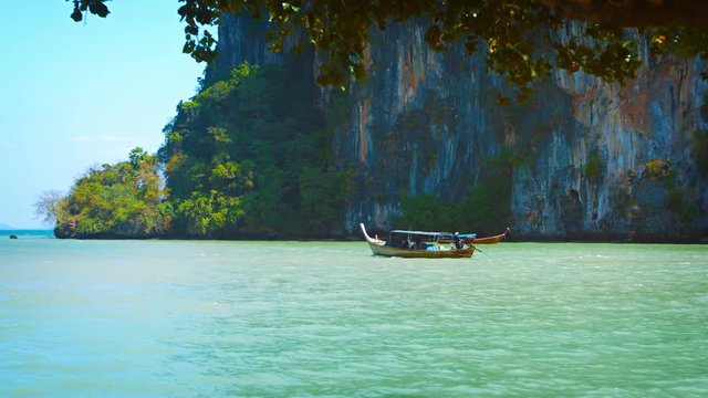 Video 2160p - Handmade, wooden longtail boat, cruising slowly along at the base of limestone seacliffs at a tropical island on a sunny day.