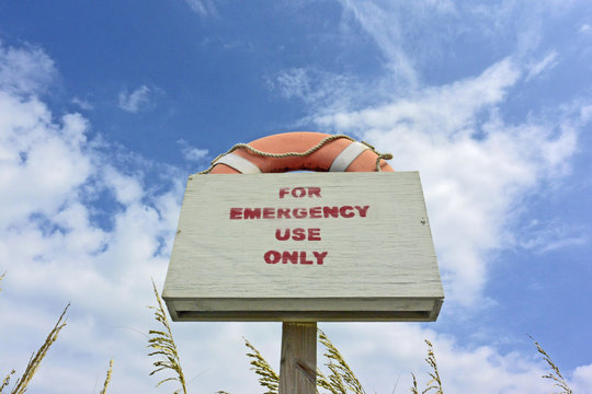 A life preserver with a sign against a blue sky with clouds and sea oats.