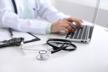 Close-up of stethoscope is lying on the table near female doctor typing on laptop computer