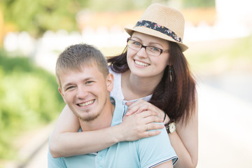 Fototapeta premium Portrait of attractive young loving couple piggybacking and smiling outdoors on a sunny summer day. A man in a blue T-shirt is carrying his woman wearing glasses and a hat.