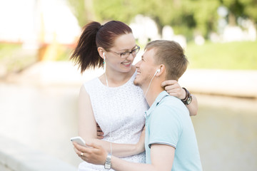 Portrait of attractive happy young couple in love, embracing, listening music on mobile phone with headphones in ears. Smiling man holding phone, woman sitting on his lap, hugging him in park