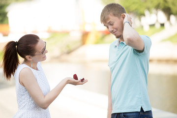 Pretty young woman requesting hand of her beloved boyfriend in park, asking to marry. Smiling girl holding box with engagement ring. Puzzled Man scratching head, thinking what to do