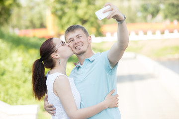 Beautiful young loving couple standing in park embracing. Pretty casual woman kissing her handsome man. He is holding a phone and making a selfie on sunny summer day