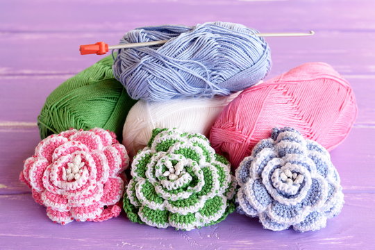 Pink, green and blue crochet roses decorated with beads. Cotton yarn skeins, hook and bright knitted flowers on wooden background lilac. Home decor crochet project